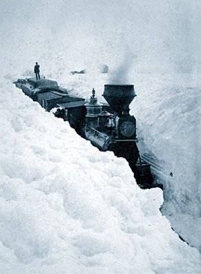 An old photo showing a train stuck in a huge snow blizzard. A man stands on one of the back carriages as the train remains stuck in the deep snow.