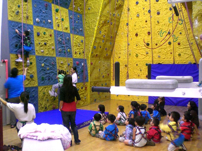This photo shows a group of children learning how to climb on a kids climbing wall. The walls are brightly colored and feature a large number of holds and possible routes to make the challenge manageable for beginners and the inexperienced.