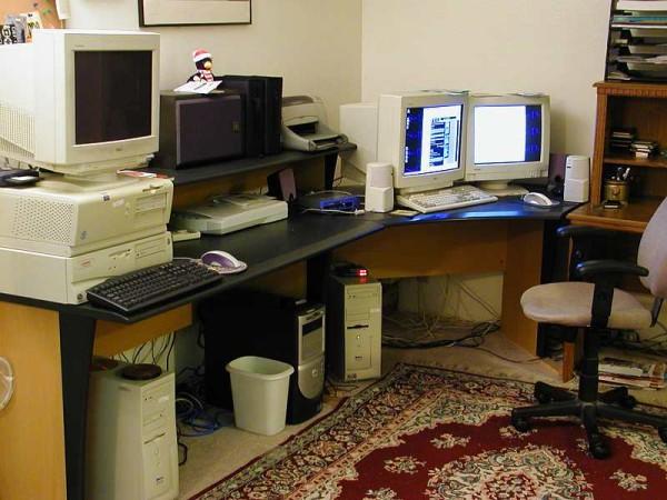 A room with a number of computers can be seen in this photo. Multiple hard drives can be seen on the ground while the monitors sit on top of the desk. There is also a chair, speakers, documents and other items in what appears to be someone's work space.
