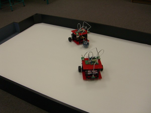 Two robots can be seen playing a game of robot soccer in an enclosed field. The wheeled robots move around the area using various sensors and algorithms to help perform better than the opponent. Touch sensors are used to avoid the walls and light sensors are used to track the ball which sends out an infrared signal.
