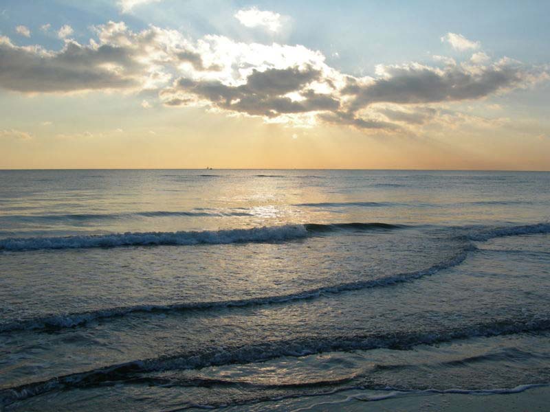A beautiful day at a beach is highlighted by the suns rays which pierce the clouds and gently illuminate the water as the small waves gently ripple on the shore. 