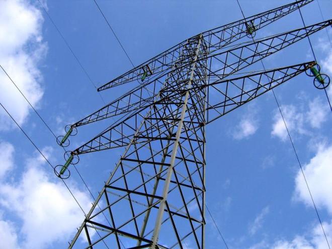 This photo looks up at the imposing structure of high voltage overhead powerlines. These structures are common all over the world as they have the important job of holding up the huge lengths of powerlines that carry electricity from the source of production to various locations where it is needed. There is a blue sky in the background with just a few scattered clouds visible.