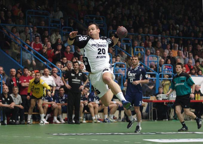 A handball player leaps high into the air as he attempts to score a goal. A large crowd can be seen watching in the photo background.