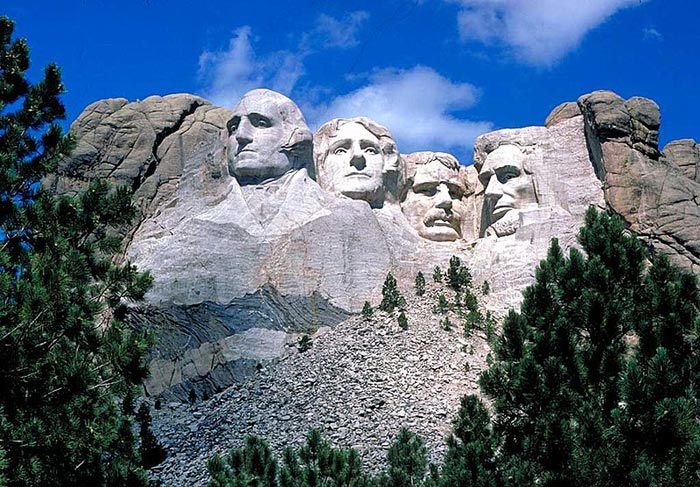 Mount Rushmore National Memorial is a huge granite sculpture that was created to celebrate 150 years of history in the United States of America. The giant sculptures are of (from left to right) George Washington, Thomas Jefferson, Theodore Roosevelt and Abraham Lincoln. This photo shows Mount Rushmore through the trees on a sunny day. It is located near Keystone, South Dakota.