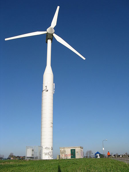 This photo shows a huge wind turbine used to harness wind power for energy. The massive structure dwarfs the person visible in the photo and stands against a beautifully blue sky background.